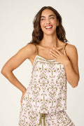 A woman with long brown hair smiles joyfully, wearing the P.J. Salvage Déjà Vu Dreams Tiki Time Camisole pajama set in pink and green, with one hand on her chest and the other on her hip.