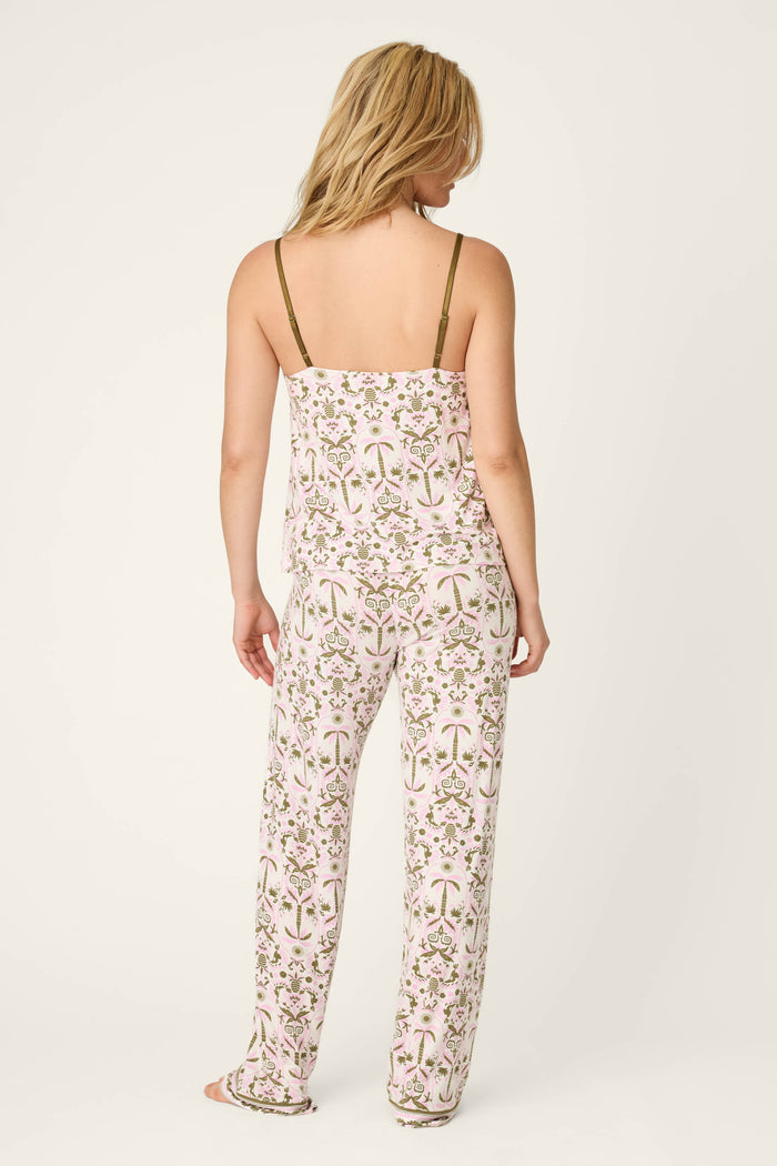 A woman with shoulder-length blonde hair stands facing away, wearing the P.J. Salvage Déjà Vu Dreams Tiki Time Long PJ Set—a white, floral pajama set featuring pink and green patterns, a spaghetti strap top, and loose-fitting pants.