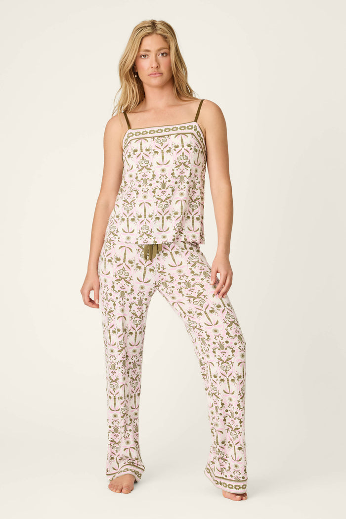 A woman stands barefoot in the P.J. Salvage Déjà Vu Dreams Tiki Time Open Leg Pants, wearing a matching floral pajama set with pink, green, and gold tones. The set features a sleeveless top with thin straps and wide-leg pants against a plain background.