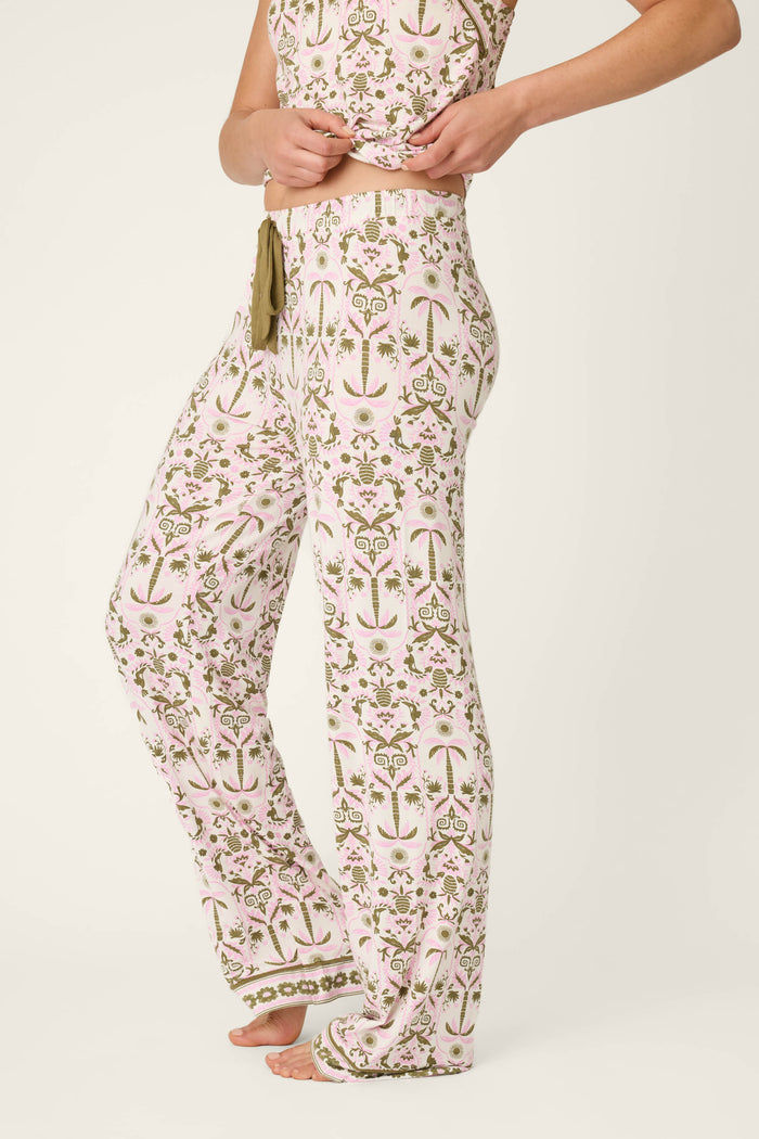 A person stands barefoot in P.J. Salvage Déjà Vu Dreams Tiki Time Open Leg Pants with a matching top, gently lifting the shirt to show the pants' waistband against a plain off-white background.