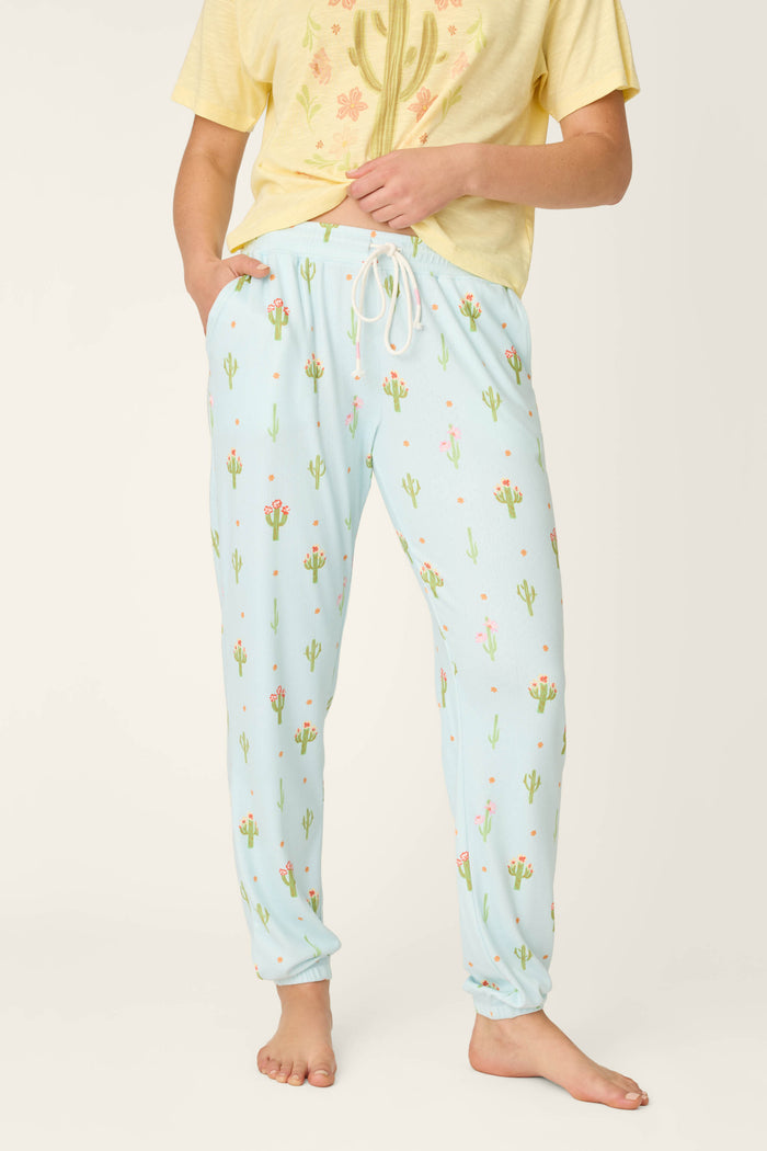 A person stands barefoot in P.J. Salvage Feelin' Prickly Cactus Blooms Banded Pants—light blue pajama pants with cactus and flower prints—and a pale yellow floral T-shirt, hands in pockets, perfect for cozy nights.