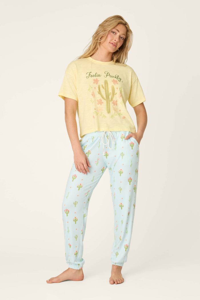 A woman stands barefoot in a yellow Feelin Prickly T-shirt with cacti and flower graphics, and P.J. Salvage Feelin' Prickly Cactus Blooms Banded Pants decorated with small cactus designs, posing against an off-white background.