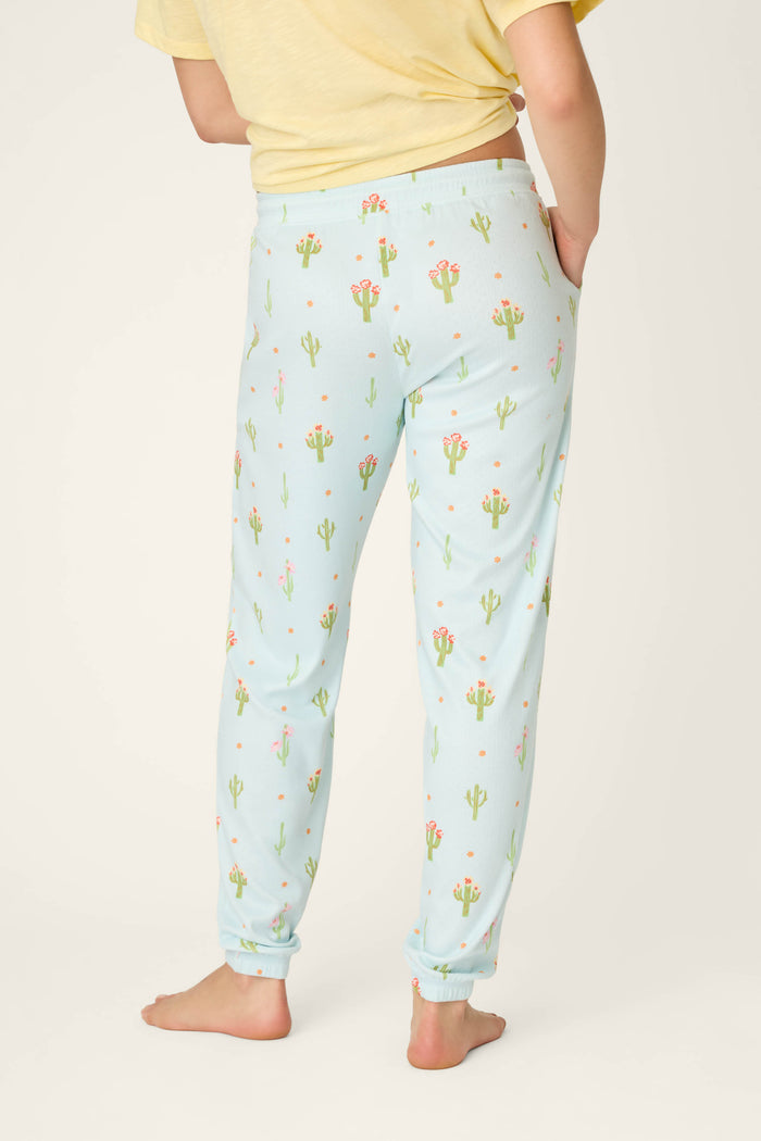 A person stands barefoot wearing P.J. Salvage Feelin' Prickly Cactus Blooms Banded Pants with a yellow T-shirt. The photo from behind shows the light blue cactus-and-flower print and relaxed fit—ideal for cozy nights.