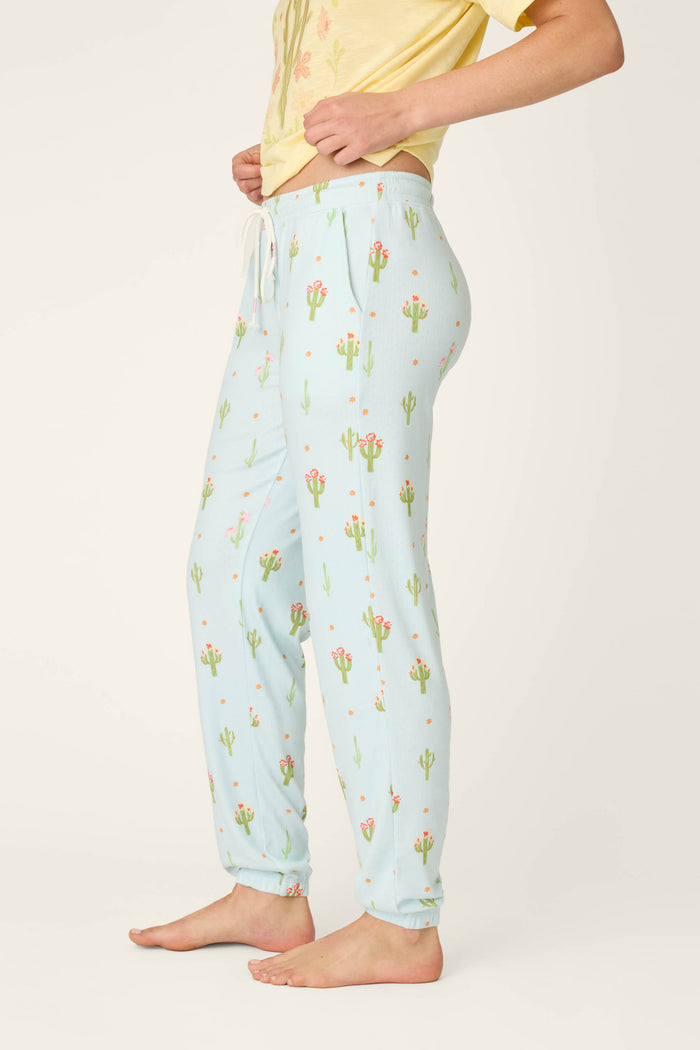 A person stands barefoot in P.J. Salvage Feelin' Prickly Cactus Blooms Banded Pants—relaxed fit, light blue with a cactus print—paired with a yellow T-shirt, shown from the side against a plain background. Perfect for cozy nights.