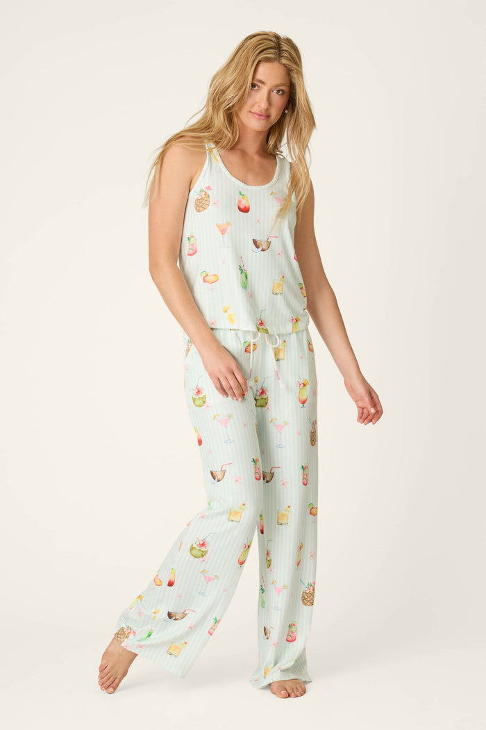 A woman with long blonde hair wears the P.J. Salvage It's Five O'clock Somewhere Cocktail Print Long PJ Set, featuring a sleeveless top and loose pants with colorful cocktail prints. She stands barefoot against a light background.