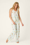 A woman with long blonde hair wears the P.J. Salvage It's Five O'clock Somewhere Cocktail Print Long PJ Set, featuring a sleeveless top and loose pants with colorful cocktail prints. She stands barefoot against a light background.
