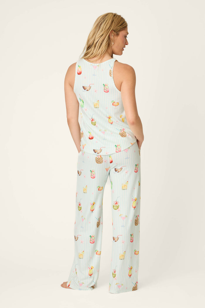 A woman stands barefoot, facing away, wearing the P.J. Salvage It's Five O'clock Somewhere Cocktail Print Long PJ Set—a sleeveless, light blue pajama set with colorful tropical drinks and subtle stripes.