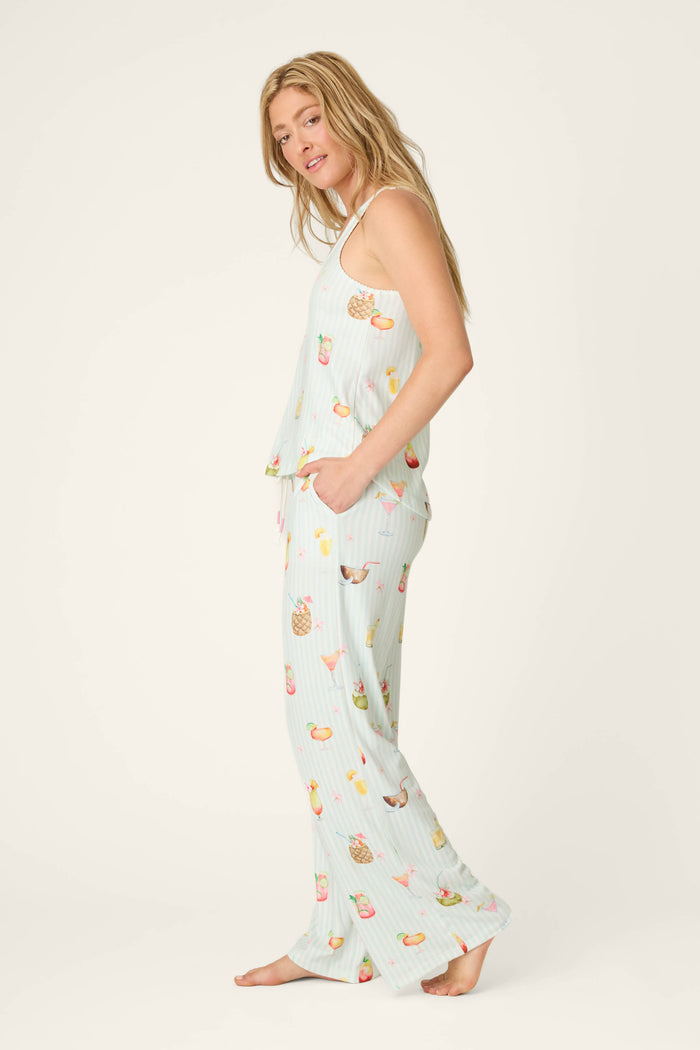 A woman stands barefoot in the P.J. Salvage It's Five O'clock Somewhere Cocktail Print Long PJ Set, featuring a sleeveless top and wide-leg pants with pockets in light blue fabric and colorful cocktail prints, facing sideways toward the camera.