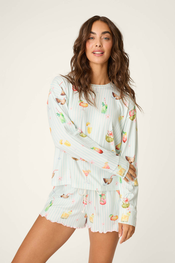 A woman with long wavy brown hair wears the P.J. Salvage "It's Five O'clock Somewhere Cocktail Print" long-sleeve top and matching shorts, featuring pastel stripes and colorful drink illustrations, standing against a plain light background, smiling slightly.