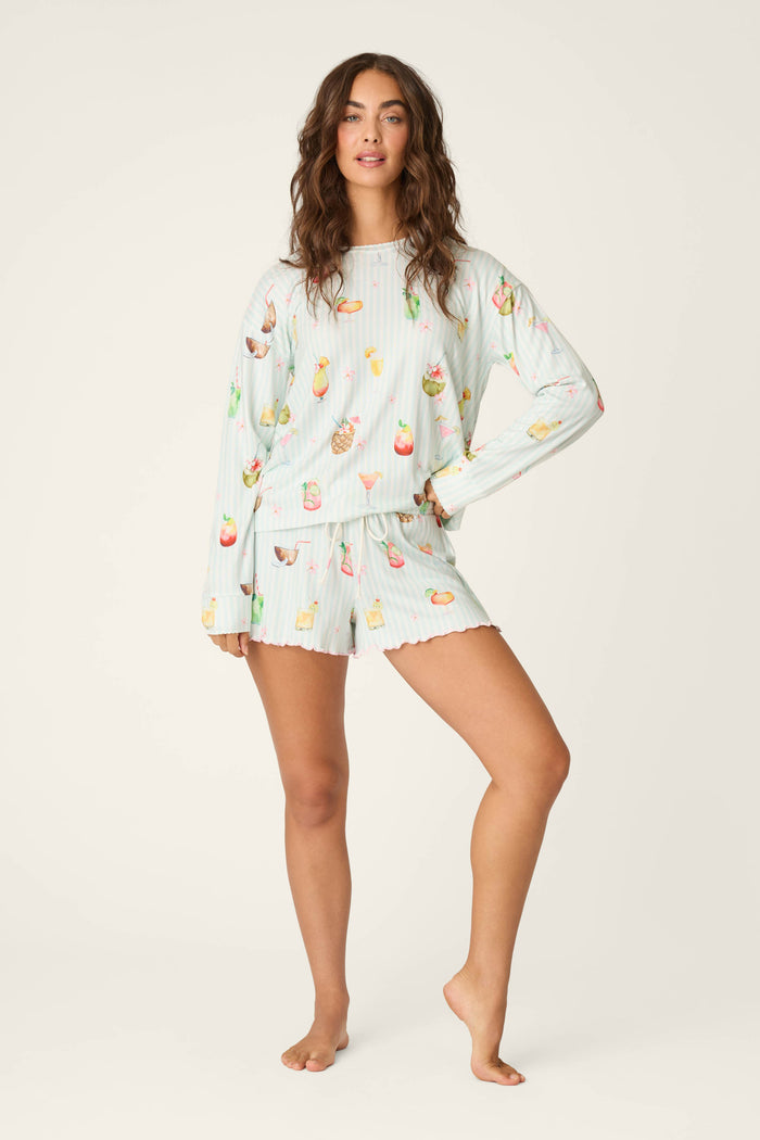 A woman with wavy hair and a cheerful look poses against a light background in P.J. Salvage's It's Five O'clock Somewhere Cocktail Print Long Sleeve Top, featuring a vibrant fruit and drink pattern.
