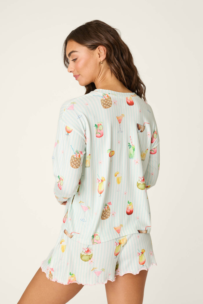A woman with long brown hair wears the P.J. Salvage "It's Five O'clock Somewhere Cocktail Print" long sleeve top, featuring colorful cocktail illustrations, as she turns to show the back against a plain background.