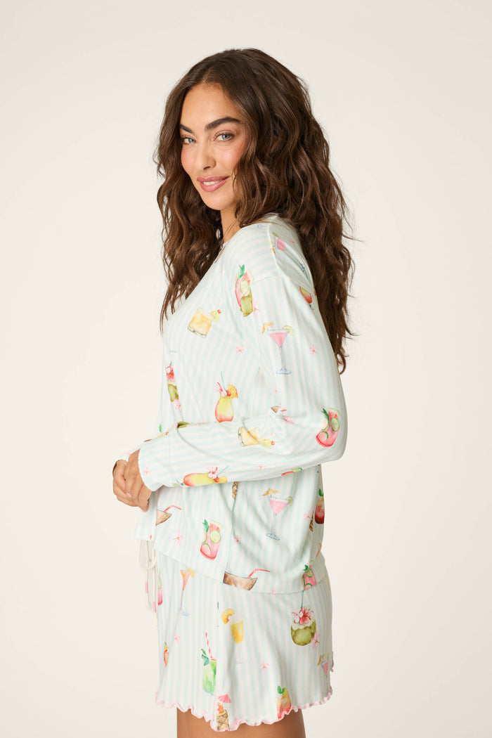 A woman with long, wavy brown hair wears the P.J. Salvage "It's Five O'clock Somewhere" Cocktail Print Long Sleeve Top, featuring colorful drink illustrations. She stands sideways and smiles gently against a plain white background.