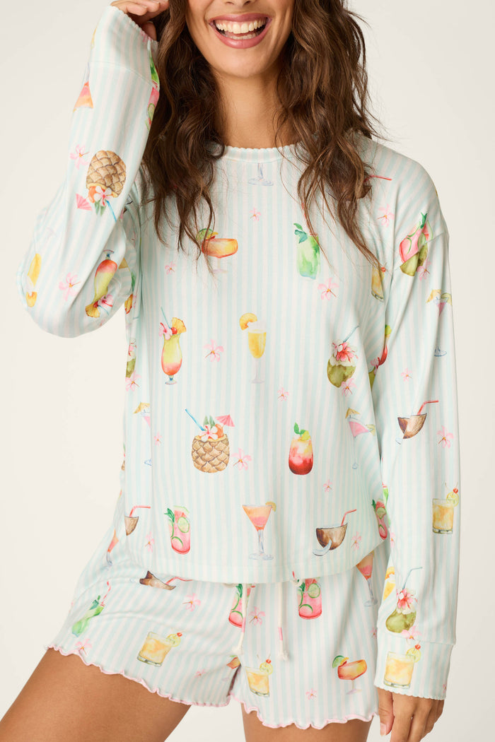 A smiling woman wears the P.J. Salvage "It's Five O'clock Somewhere" Long Sleeve Top featuring a colorful cocktail print, paired with matching striped shorts with ruffled hems.
