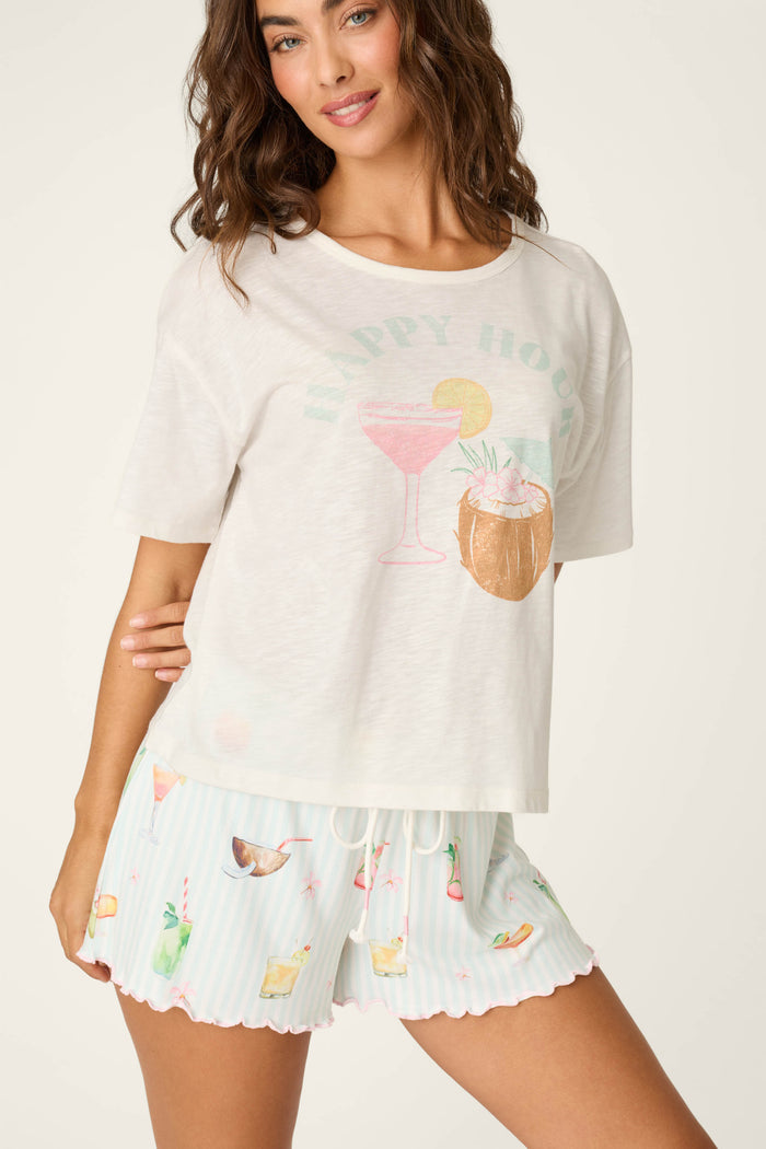 A woman smiles, posing in the P.J. Salvage It's Five O'clock Somewhere "Happy Hour" Graphic T-Shirt and light blue striped pajama shorts with colorful drink prints.