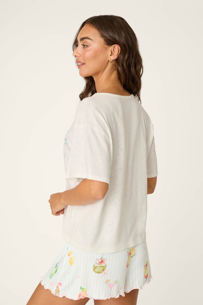 A woman with wavy brown hair smiles in profile, wearing the P.J. Salvage It's Five O'clock Somewhere "Happy Hour" Graphic T-Shirt and pastel fruit-and-flamingo print shorts, standing against a plain light background.
