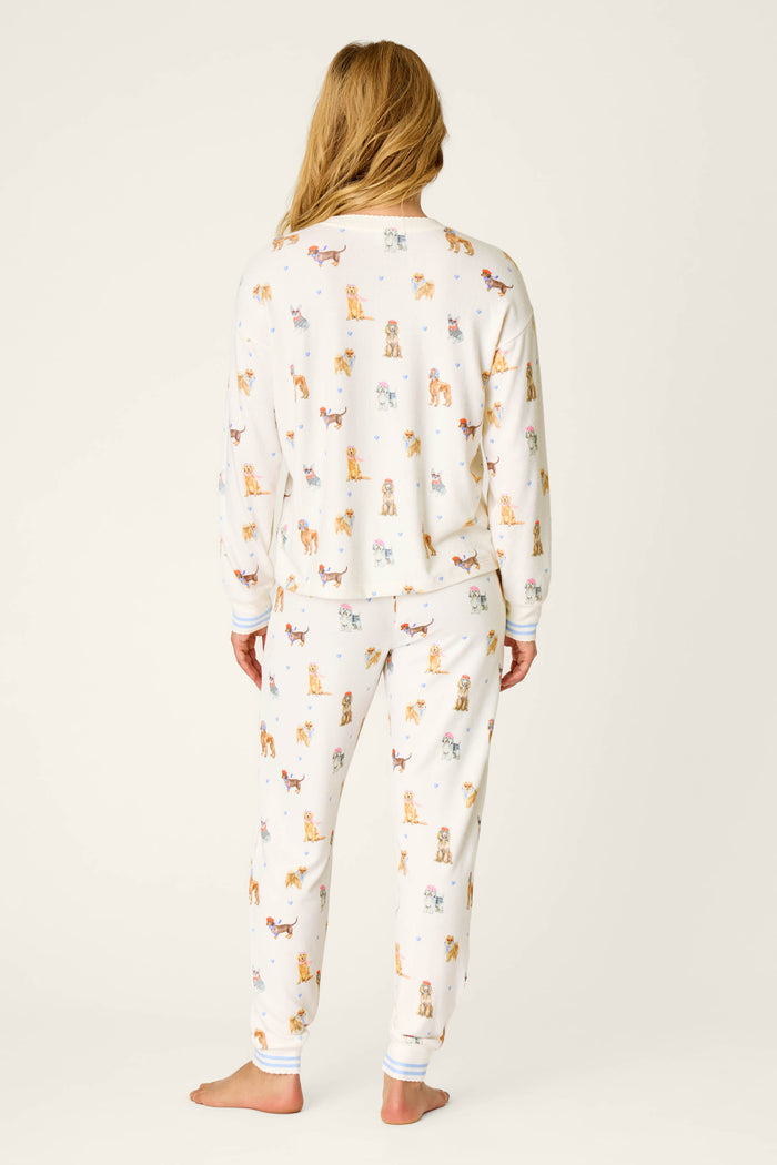 A person stands barefoot facing away, wearing the P.J. Salvage Meet Me in Pawris Dog Print Long Sleeve PJ Set, which features a white background with colorful dog prints. The pajama set includes long sleeves and pants, and is shown against a plain light-colored backdrop.