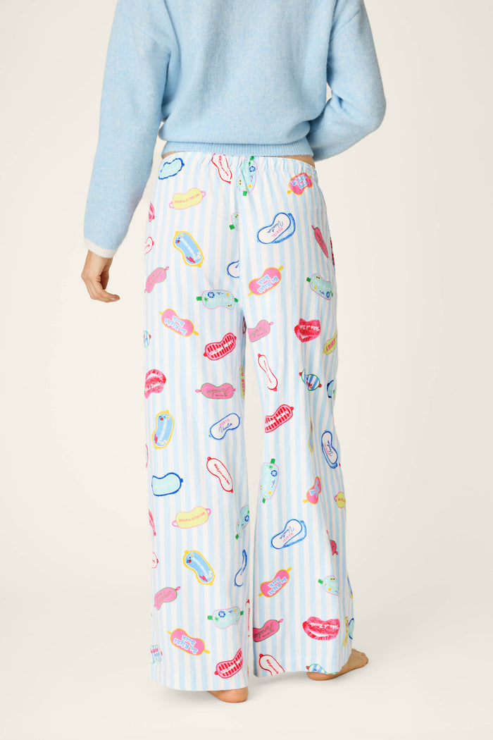 A person stands barefoot and facing away from the camera against a neutral background, wearing a blue sweater paired with P.J. Salvage's Out of Office Eyemask Print Flannel Pajama Pants—loose white pajama pants featuring colorful lip, eye, and sandal patterns.