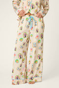 Model showcases the P.J. Salvage Picture Purrfect Pet Portraits Satin Pajama Pants, featuring a cream base adorned with vibrant cartoon dog faces and finished with a blue drawstring, standing barefoot against a light background. Only the lower half of the body is visible.
