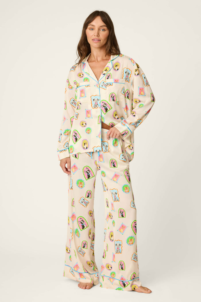 A woman stands barefoot in P.J. Salvage's Picture Purrfect Pet Portraits Satin Pajama Pants, featuring loose, light-colored fabric adorned with playful, colorful cartoon pet faces and objects. She looks forward with a gentle smile against a plain background.