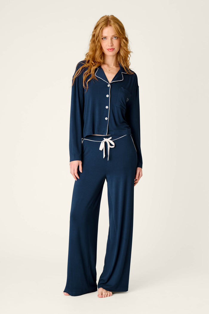 A woman with long red hair wears the Soiree Dreams Deep Navy Long Sleeve PJ Set by P.J. Salvage, featuring white trim and buttons, as she stands barefoot against a plain light background.